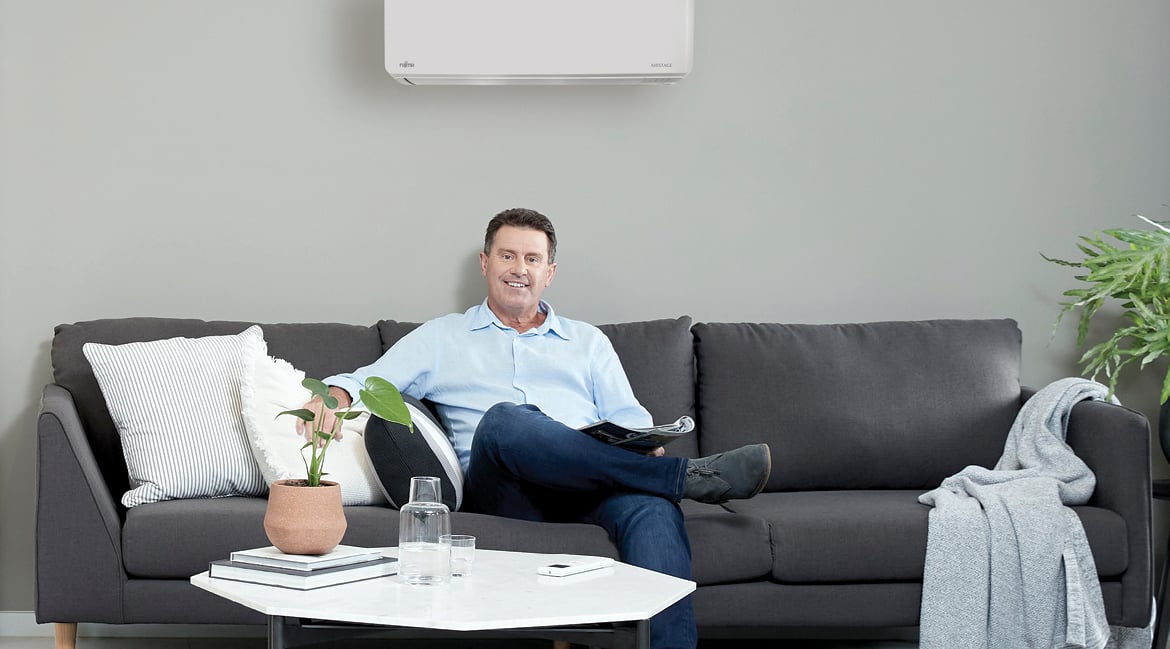 A man reading on a grey couch with a Fujitsu wall split conditioner above — quiet, steady comfort in the background.